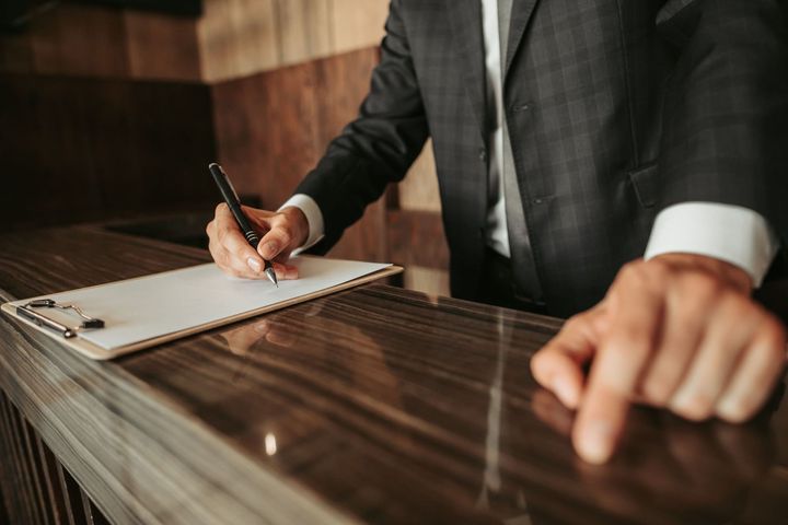 Hombre con traje escribiendo en un portapapeles en un mostrador, probablemente en una oficina o un hotel.