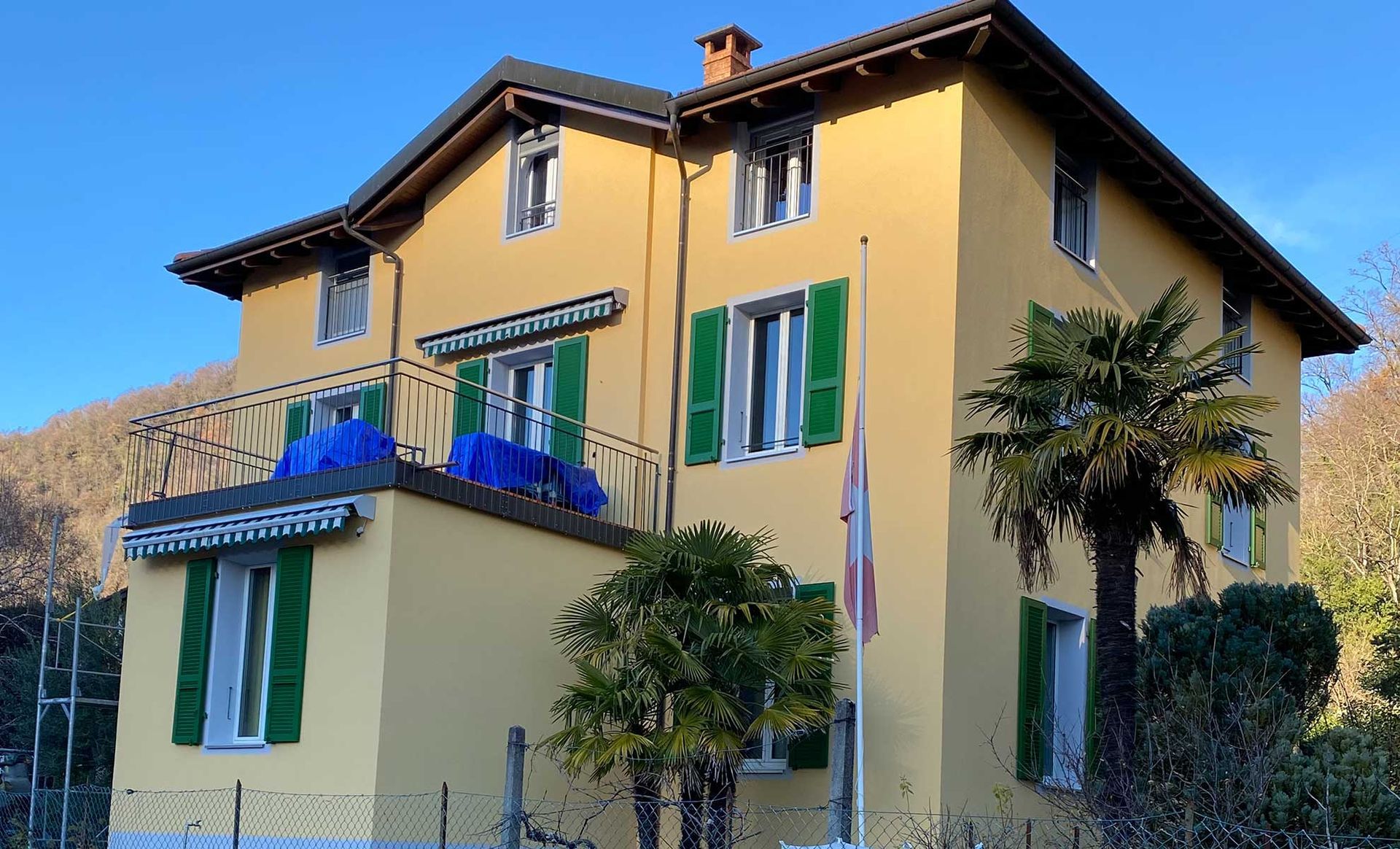 Edificio giallo con persiane verdi, una bandiera svizzera e una palma in una giornata di sole.