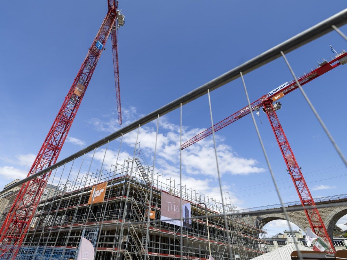 Cantiere con gru rosse contro il cielo azzurro, visto attraverso una recinzione metallica.