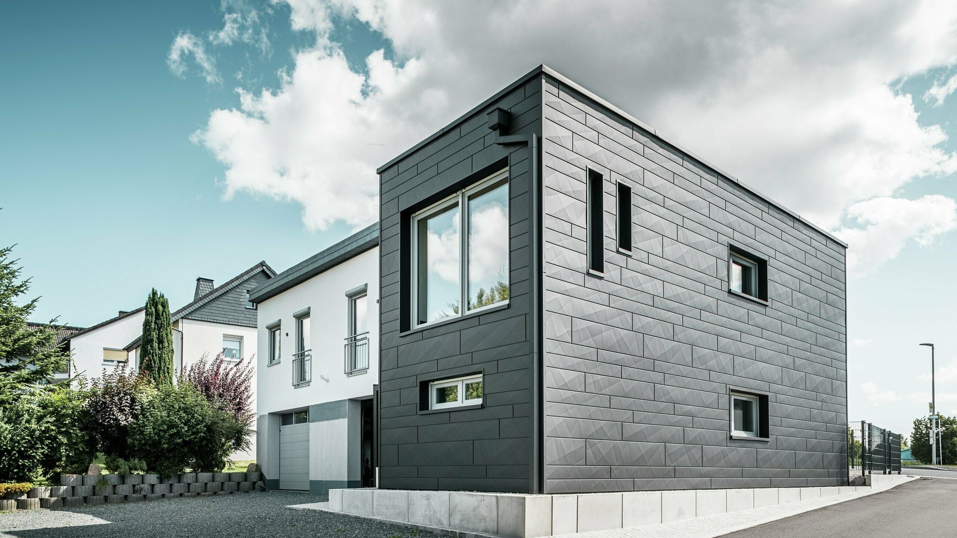 Moderna casa nera con facciata grigia accanto a una casa bianca con cielo azzurro.
