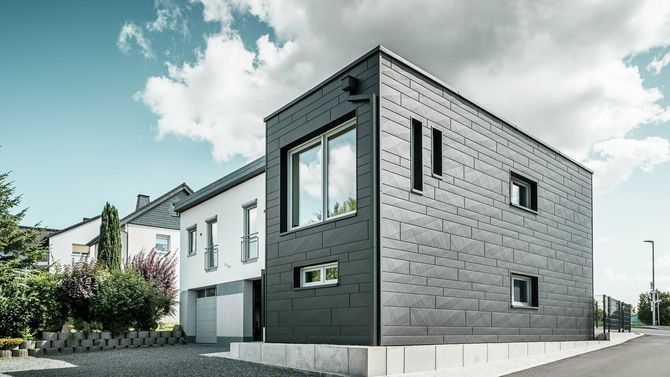 Moderna casa nera con facciata grigia accanto a una casa bianca con cielo azzurro.
