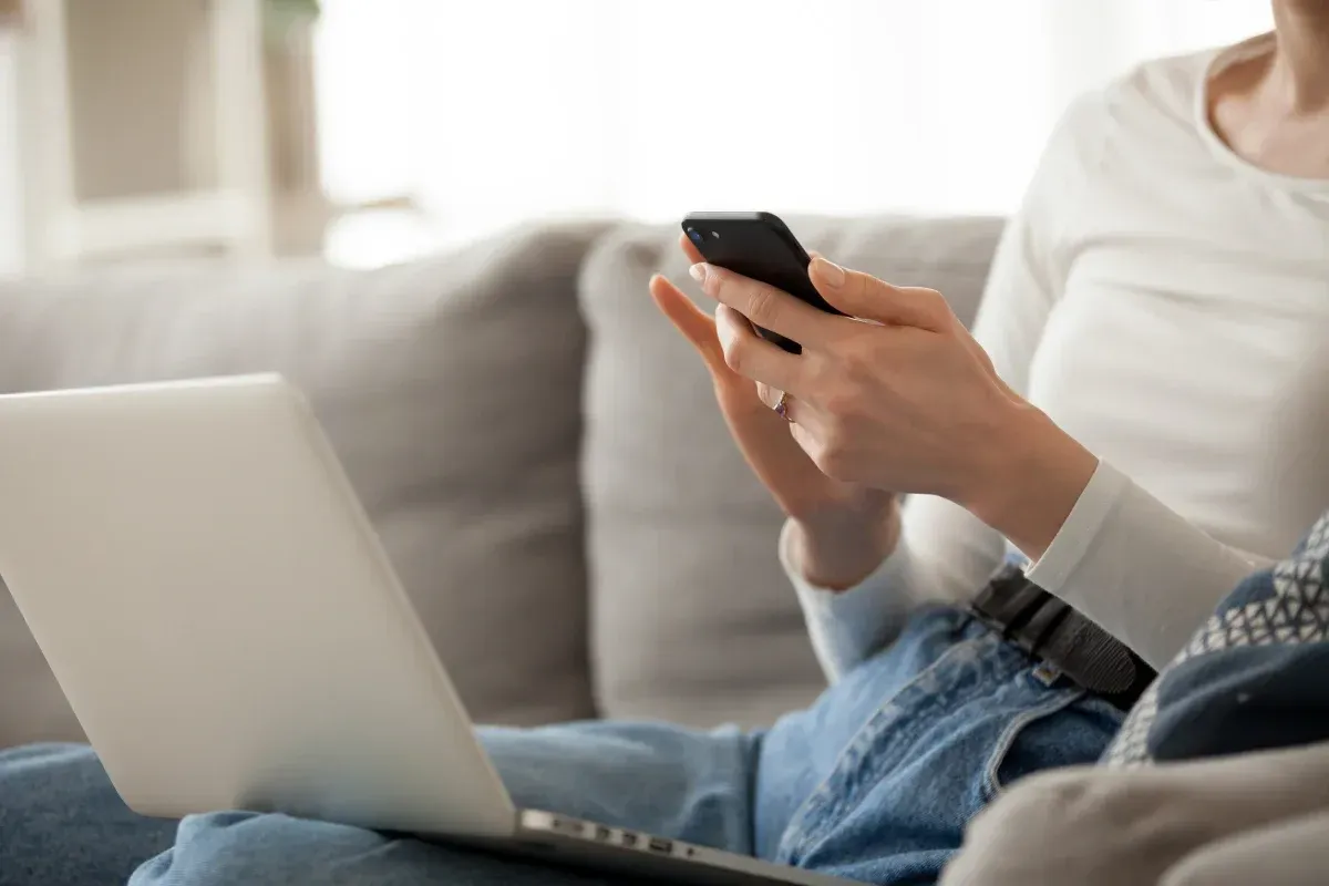 Persona sentada en un sofá usando un teléfono inteligente y una computadora portátil.