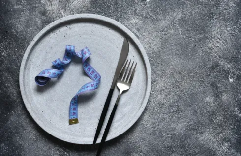 Plato gris con una cinta métrica azul, un cuchillo y un tenedor, sobre un fondo gris.