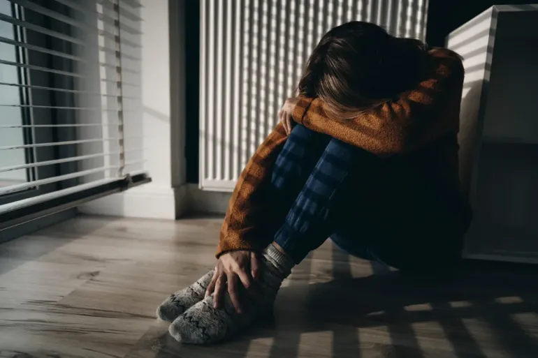 Persona acurrucada en el suelo, con los brazos rodeando las rodillas. Las persianas proyectan franjas de luz.