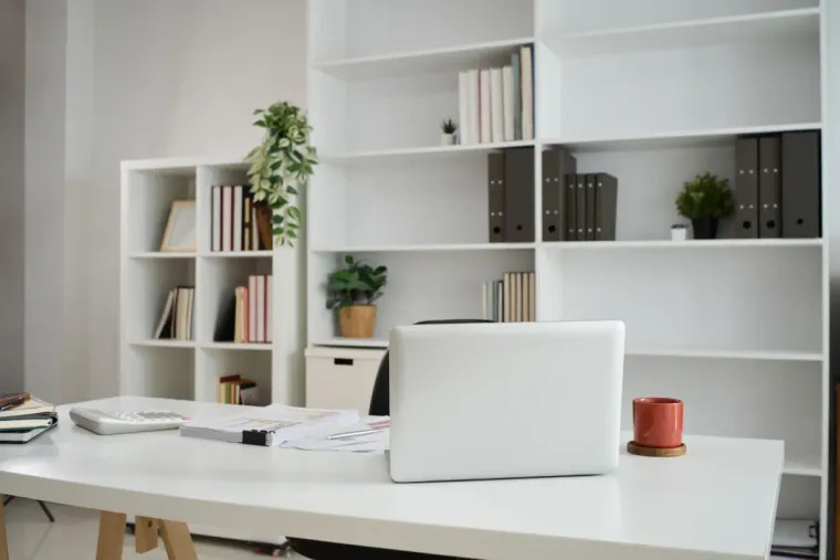 Espacio de trabajo con escritorio negro, estantes y silla azul verdosa; computadora portátil abierta, ventana con persianas.