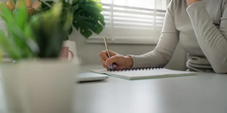 Una persona escribe en un cuaderno de espiral sobre un escritorio blanco con plantas al fondo.
