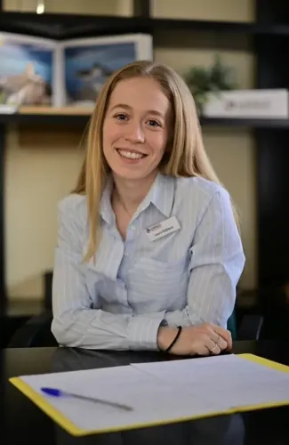 Mujer sonriente, vestida con una camisa azul claro abotonada, sentada detrás de un escritorio con papeles y un bolígrafo.