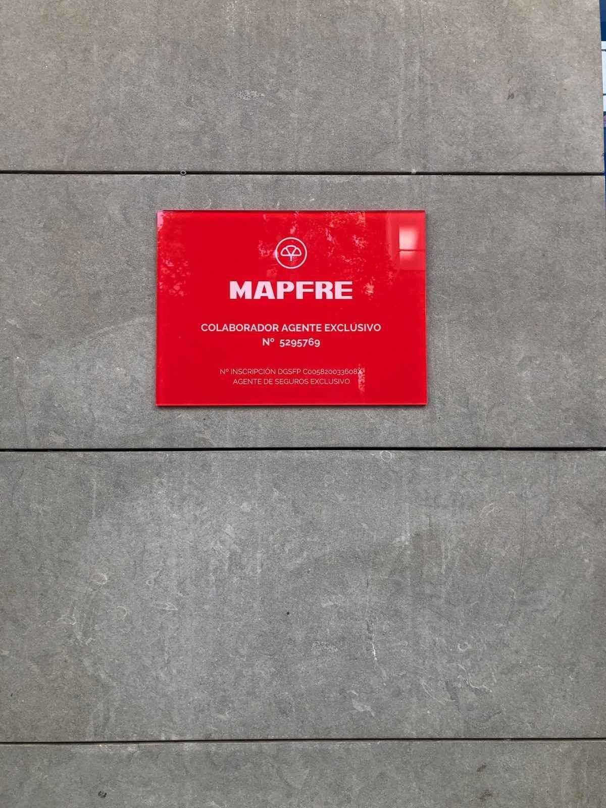 Un cartel rectangular rojo de la compañía de seguros Mapfre con texto blanco, montado sobre una pared de piedra gris.