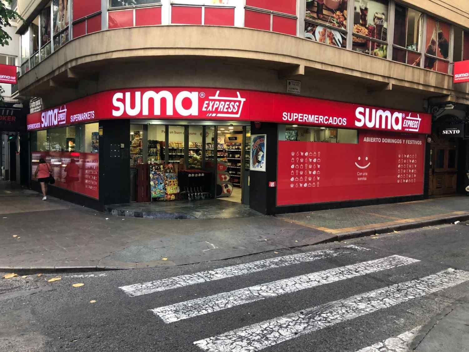 Vista desde una esquina de un supermercado rojo Suma Express en una calle de la ciudad, con un paso de peatones en primer plano.
