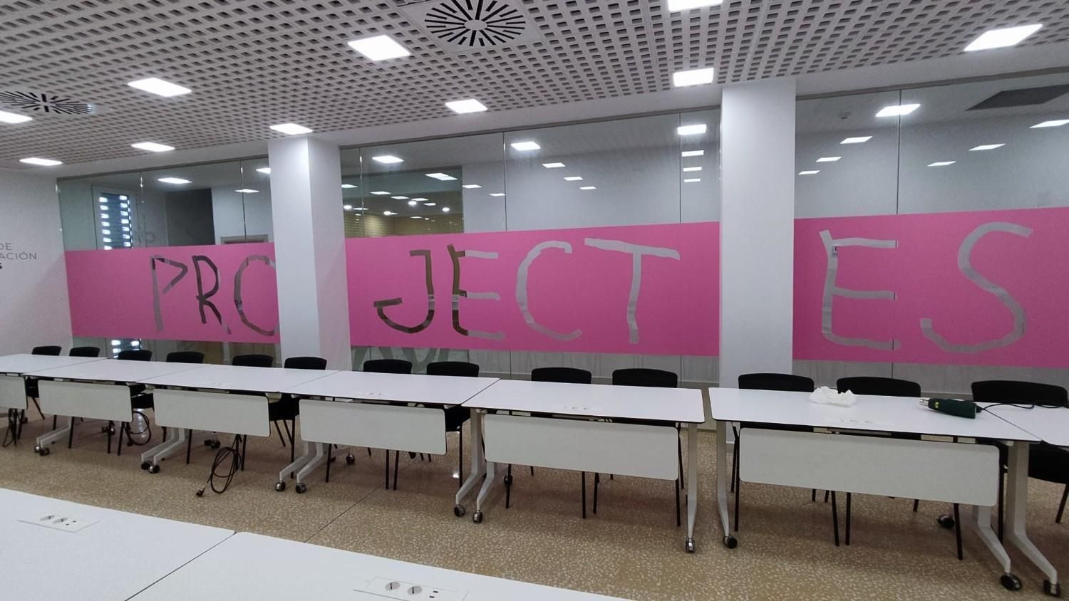 Una larga fila de mesas y sillas negras frente a una pared con una franja rosa que dice 