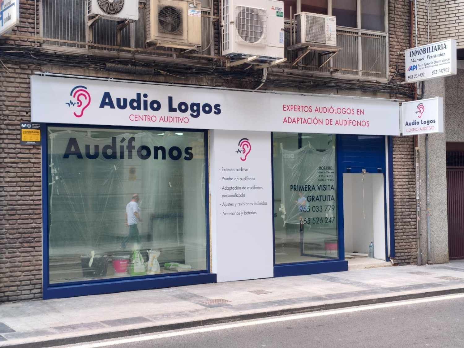 La fachada de Audio Logos, un centro de audífonos, con un letrero blanco, ventanas de cristal y una persona visible en el interior.