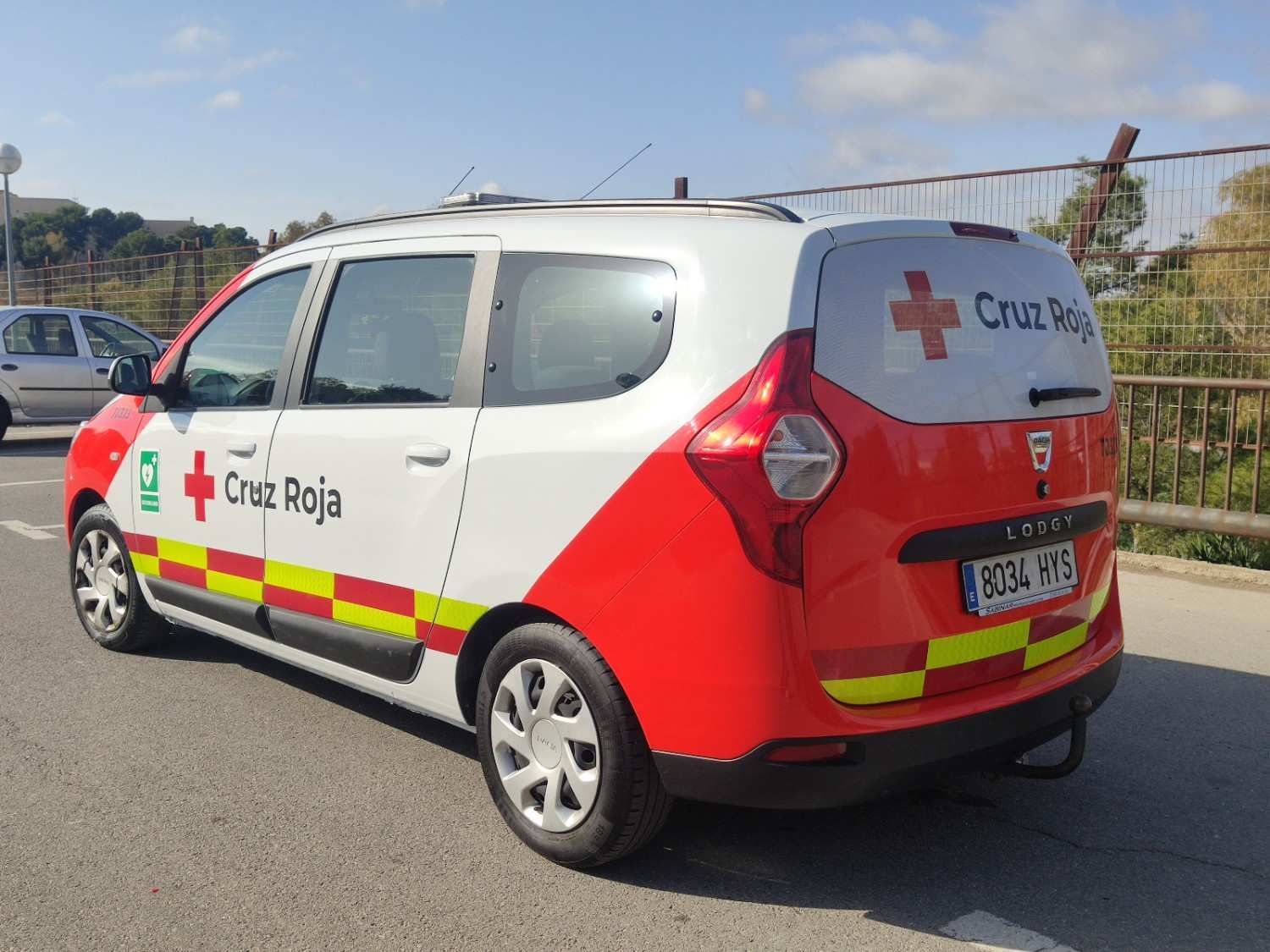 Una furgoneta de la Cruz Roja, de color blanco y rojo, con distintivos reflectantes amarillos, estaba aparcada en una carretera asfaltada.