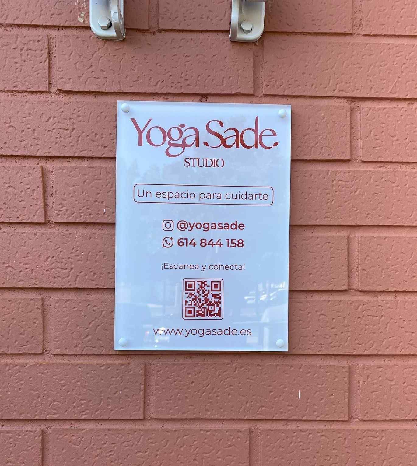 Un letrero del estudio Yoga Sade, colocado en una pared de ladrillos, con información de contacto, redes sociales y un código QR.