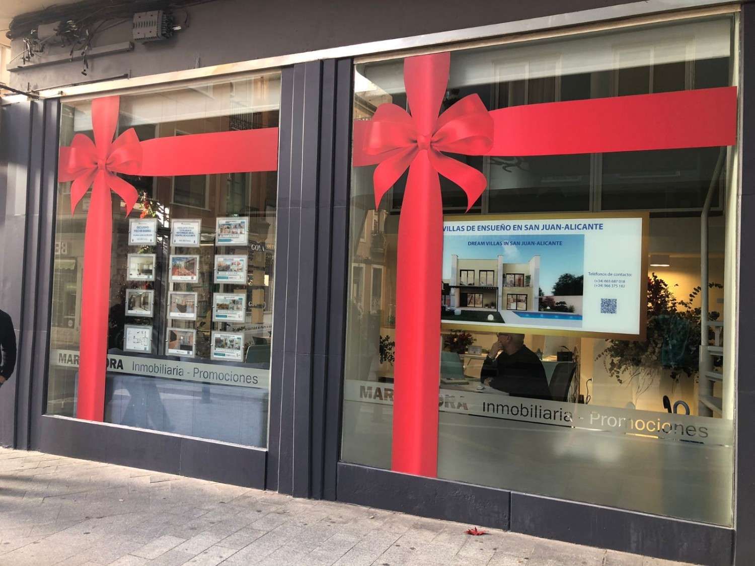 Un escaparate decorado con grandes lazos rojos como si fuera un regalo, donde se exhiben anuncios inmobiliarios y una oficina en el interior.