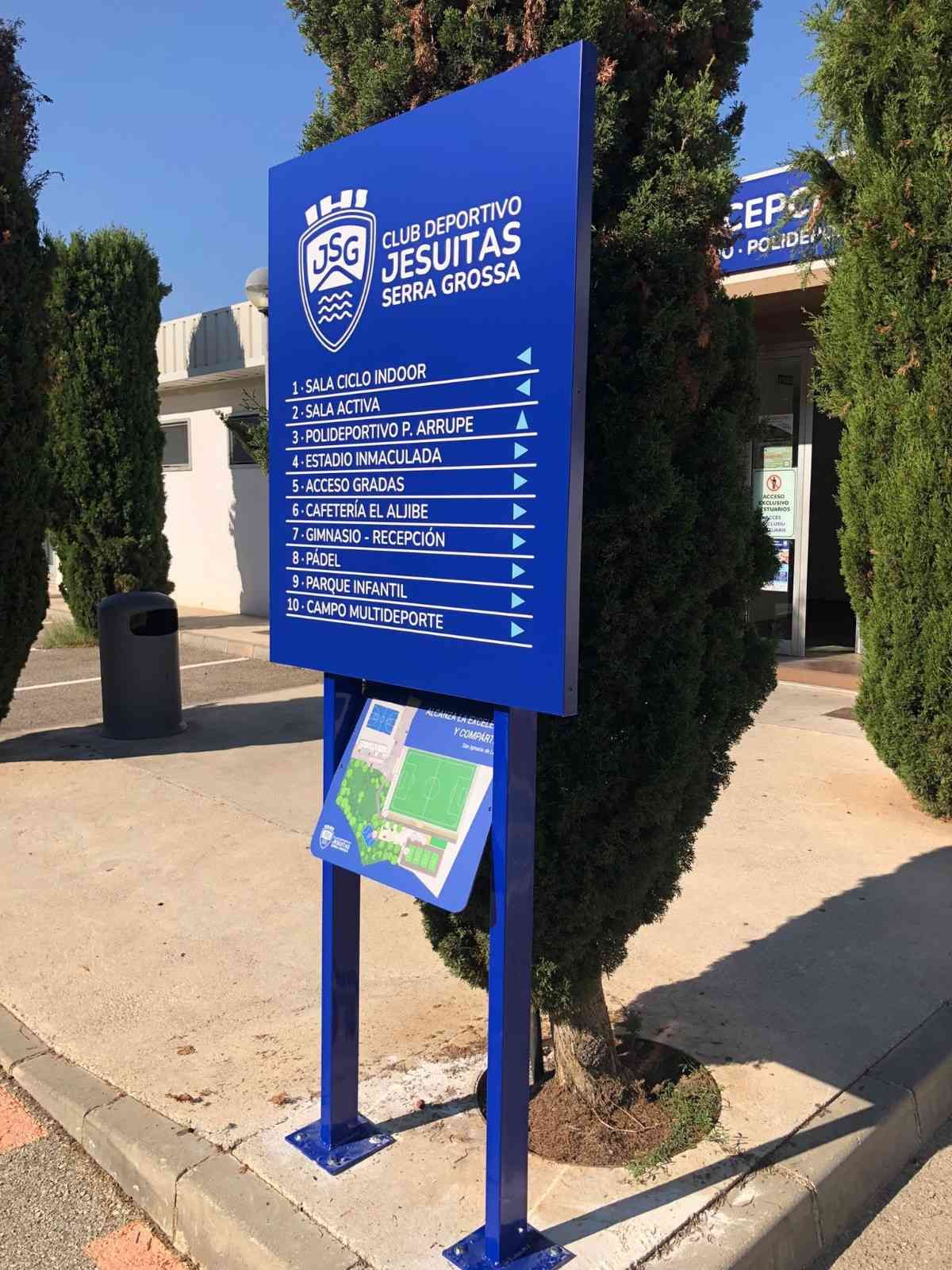 Señal azul que indica la ubicación del Club Deportivo Jesuitas, frente a un árbol, con una lista de instalaciones y un pequeño mapa de la zona.