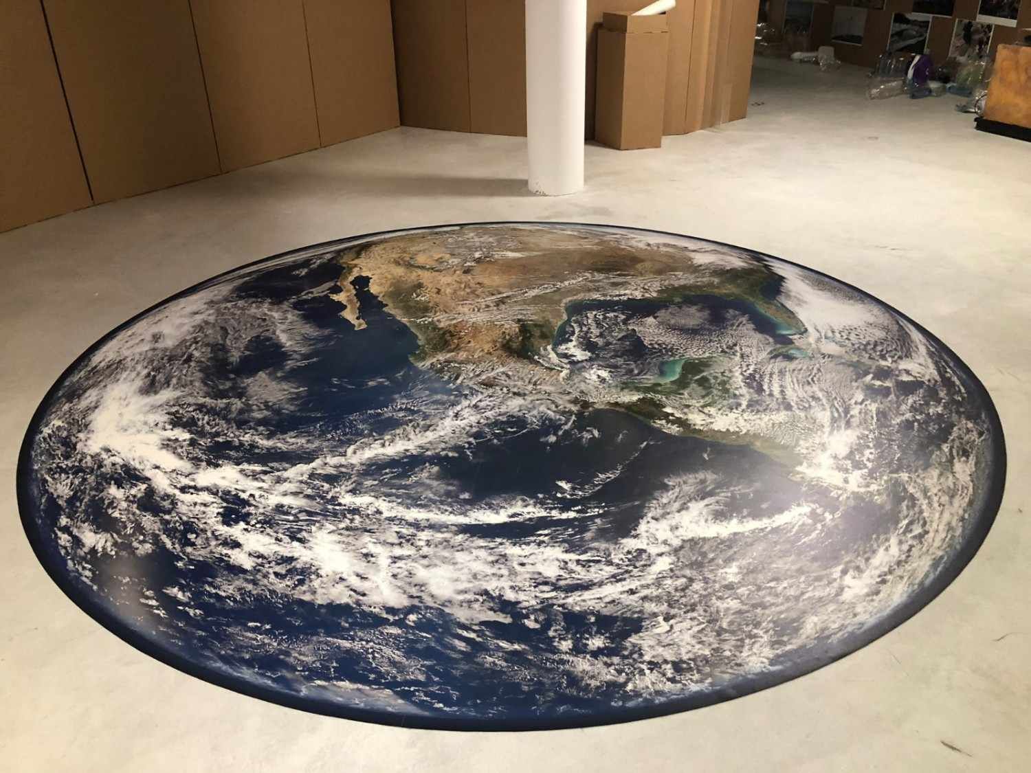 Un gran vinilo decorativo circular para el suelo que representa la Tierra vista desde el espacio, centrado sobre un suelo de color claro en un entorno similar a un almacén.