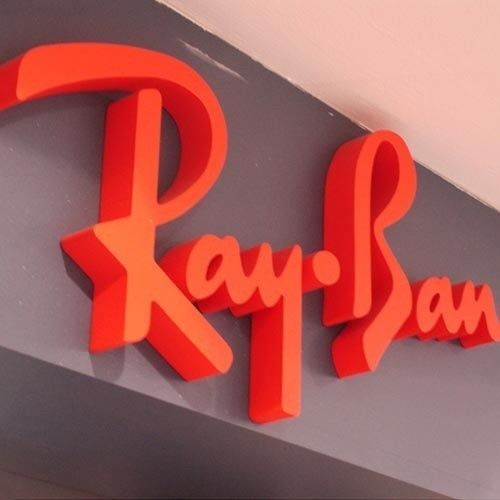 Un logotipo de Ray-Ban en 3D, de color rojo brillante, montado sobre una pared gris oscuro, visto desde un ángulo ligeramente elevado.