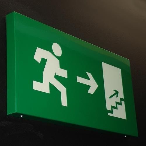 Señal de salida de emergencia de color verde con una figura blanca corriendo hacia una puerta con escaleras que indican una dirección ascendente.