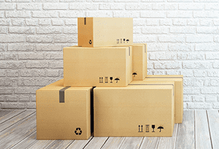 Cajas de cartón apiladas en una habitación luminosa con una pared de ladrillo blanco y suelo de madera.