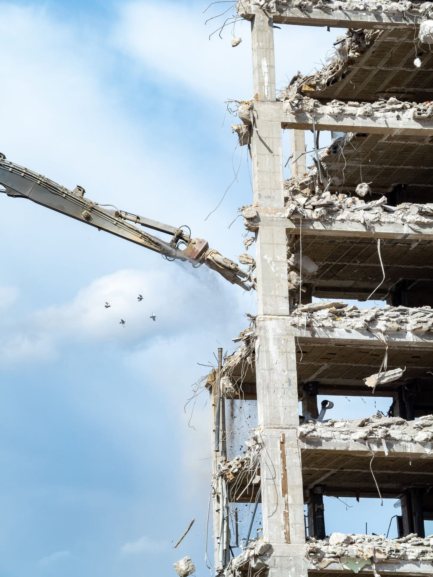 Una grúa de construcción con un brazo largo demuele los pisos superiores de un edificio de hormigón contra un cielo azul brillante.