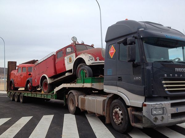 Un camión semirremolque Iveco de color gris oscuro remolcando un remolque de plataforma cargado con dos camiones de bomberos rojos antiguos en una carretera asfaltada.