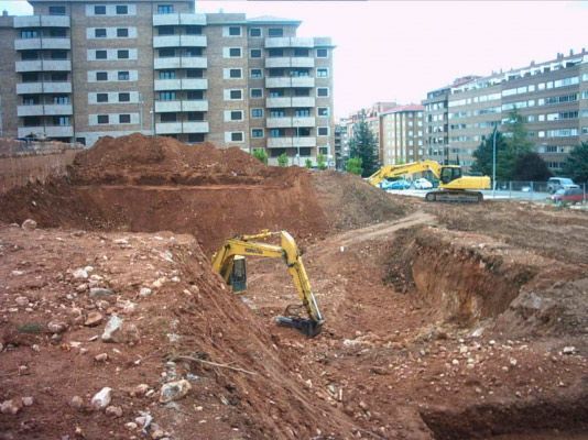 Una excavadora amarilla trabaja en una profunda fosa de tierra fangosa en una obra de construcción cerca de edificios de apartamentos urbanos.
