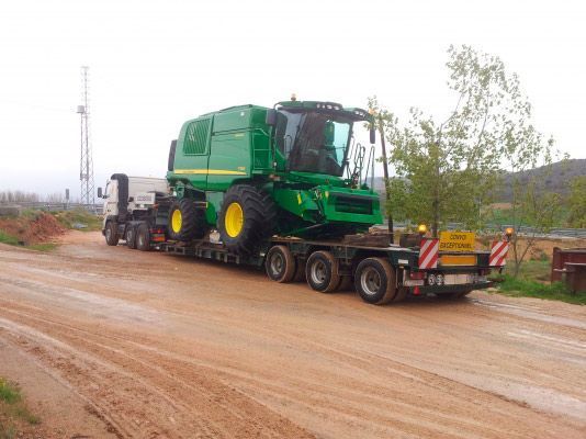 Una gran cosechadora John Deere de color verde está siendo transportada en un remolque de plataforma por un camino de tierra.