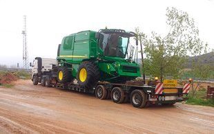 Una cosechadora John Deere de color verde siendo transportada en un remolque de plataforma plana por un camino de tierra.