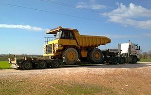 Un gran camión volquete amarillo cargado sobre un remolque de plataforma es remolcado por un camión semirremolque blanco en un camino de tierra bajo un cielo azul.
