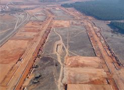 Vista aérea de una extensa zona de construcción despejada, con caminos de tierra, terreno nivelado y un bosque cercano.