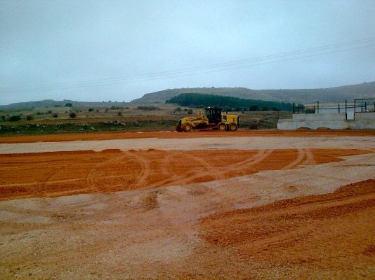 Una niveladora de construcción amarilla trabaja en un gran terreno despejado de tierra roja bajo un cielo nublado.