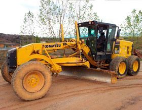Una motoniveladora Komatsu amarilla estacionada en un terreno de tierra, equipada con una gran pala central para nivelar el suelo.