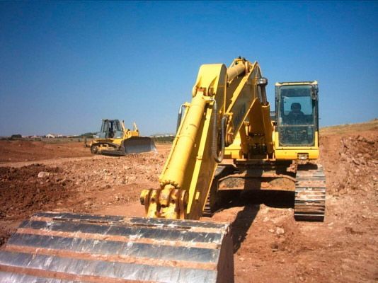 Una excavadora amarilla se encuentra en un campo de tierra bajo un cielo azul despejado, con una topadora trabajando al fondo.