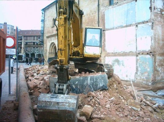 Una excavadora amarilla se encuentra sobre un montón de escombros de ladrillo mientras demuele la pared de un edificio en una calle de la ciudad.