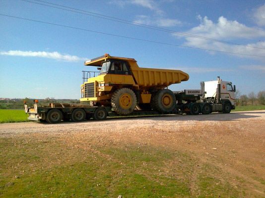 Un camión semirremolque blanco transporta un gran camión volquete minero amarillo sobre un remolque de plataforma a lo largo de un camino de grava en un campo.