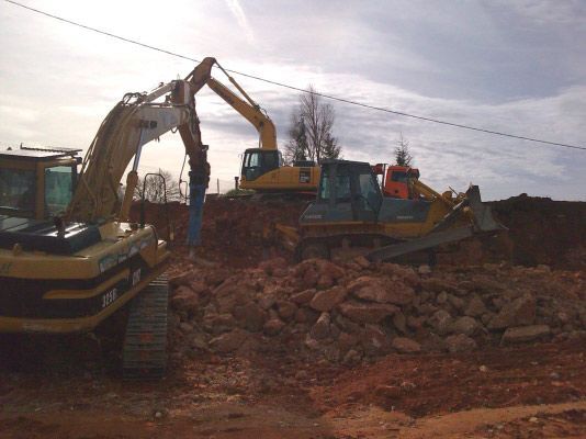 Dos excavadoras amarillas rompen rocas y remueven tierra en una obra de construcción bajo un cielo nublado.
