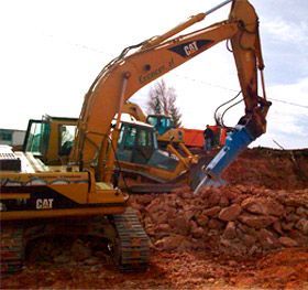 Una excavadora CAT amarilla con un martillo hidráulico incorporado está rompiendo rocas en una obra de construcción.