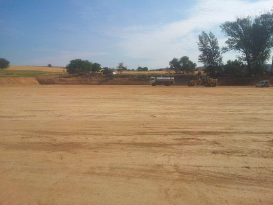 Un amplio solar llano de tierra, como una obra en construcción, bajo un cielo azul despejado, con maquinaria pesada aparcada cerca de un montículo de tierra a lo lejos.