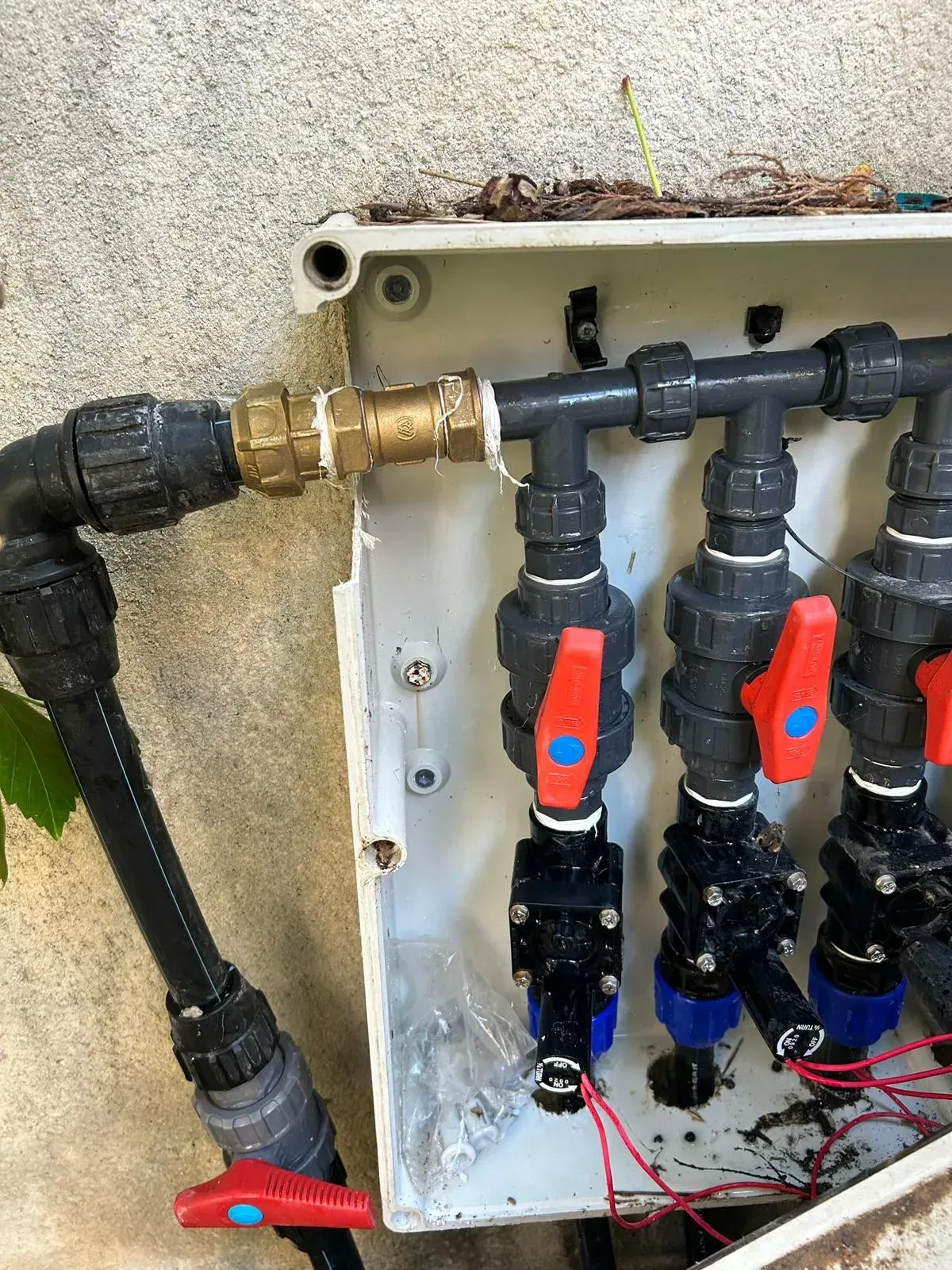 Sistema de riego con tuberías negras, válvulas y caja de control adosado a una pared desgastada.