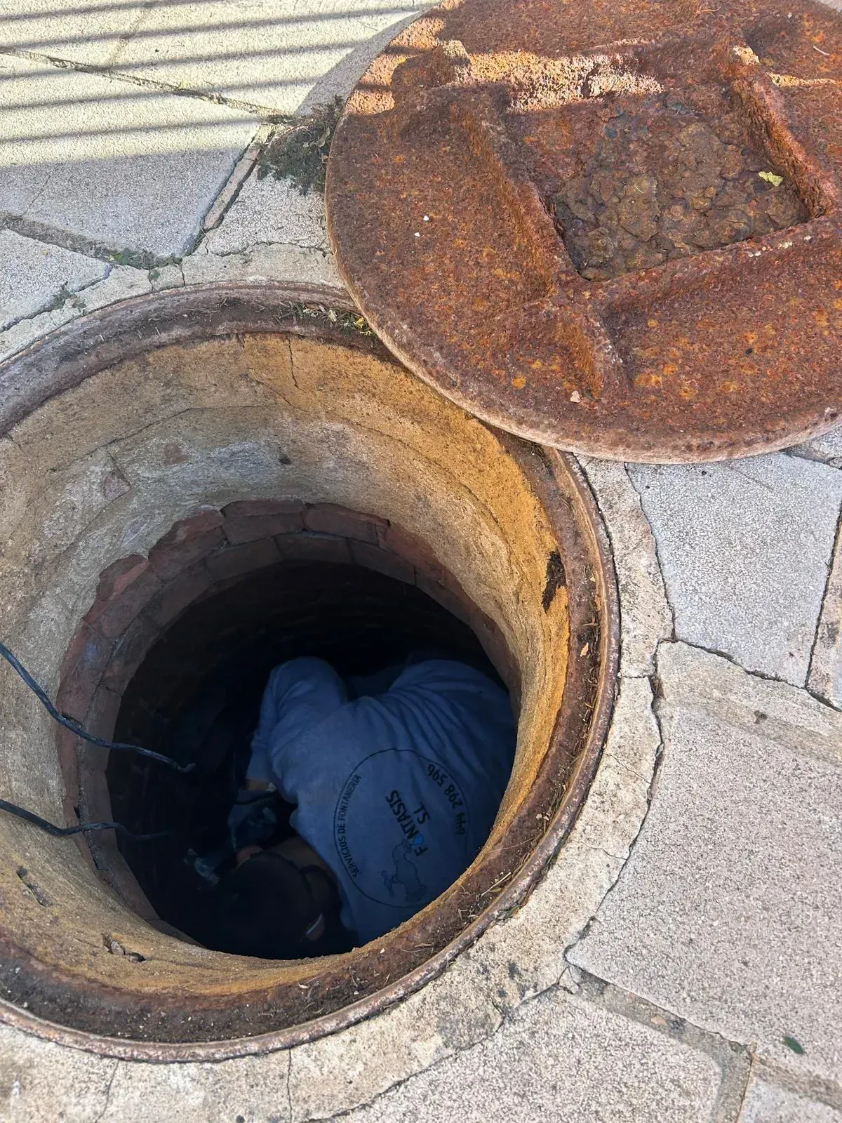 Persona dentro de una alcantarilla abierta, con la tapa entreabierta, sobre una acera de piedra.