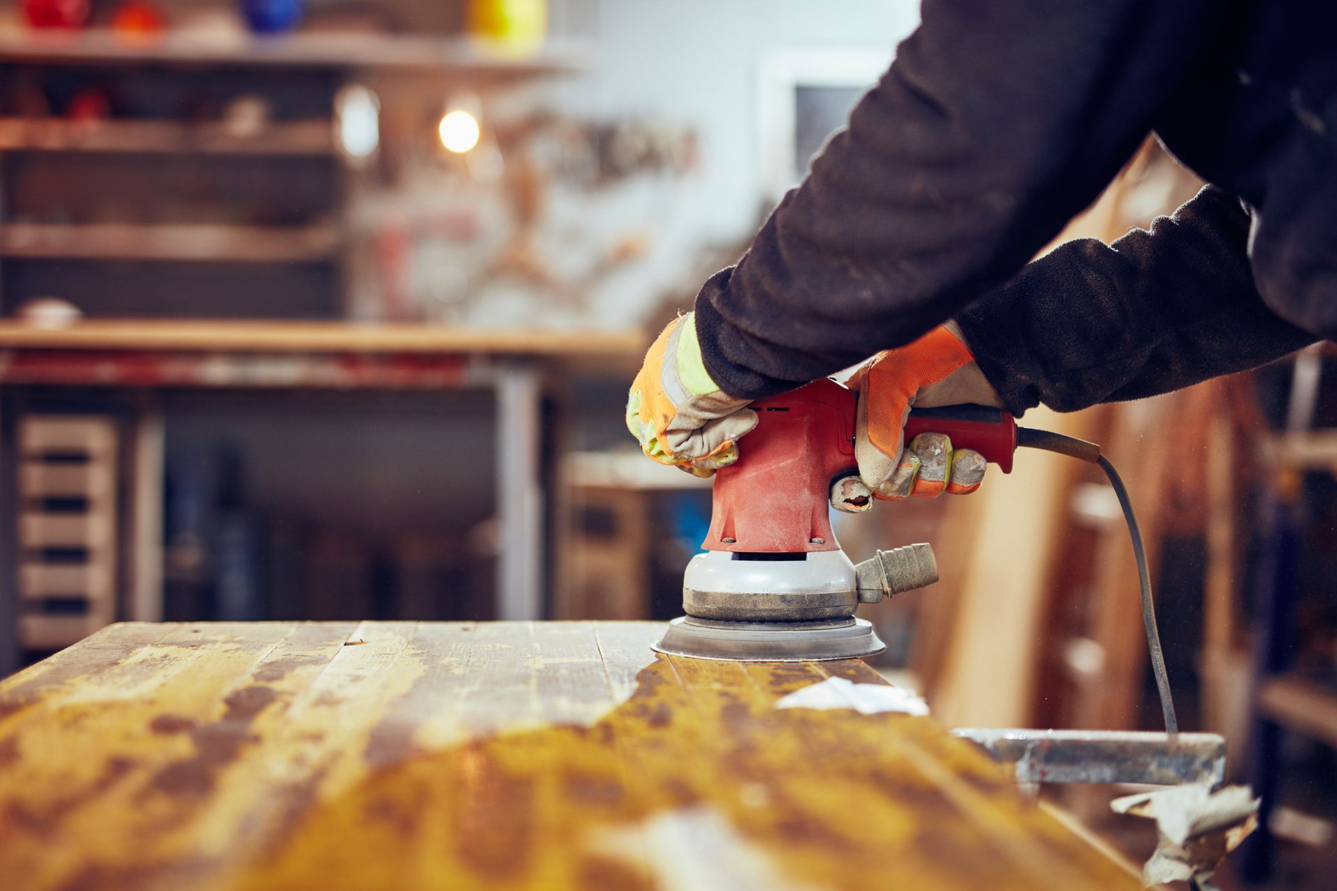 Eine Person, die Arbeitshandschuhe trägt, glättet in einer Werkstatt mit einem roten Elektroschleifer die Oberfläche eines Holzbretts.