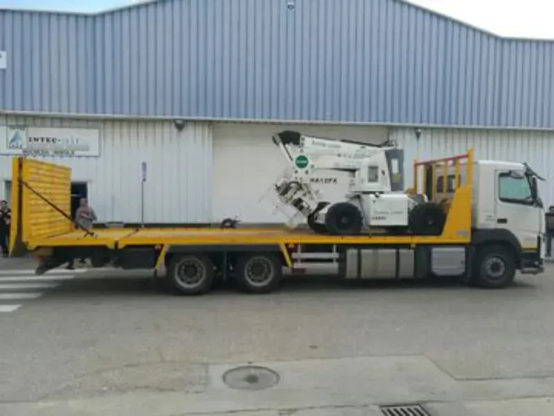 Camión de plataforma amarillo que transporta una plataforma elevadora blanca frente a un almacén.