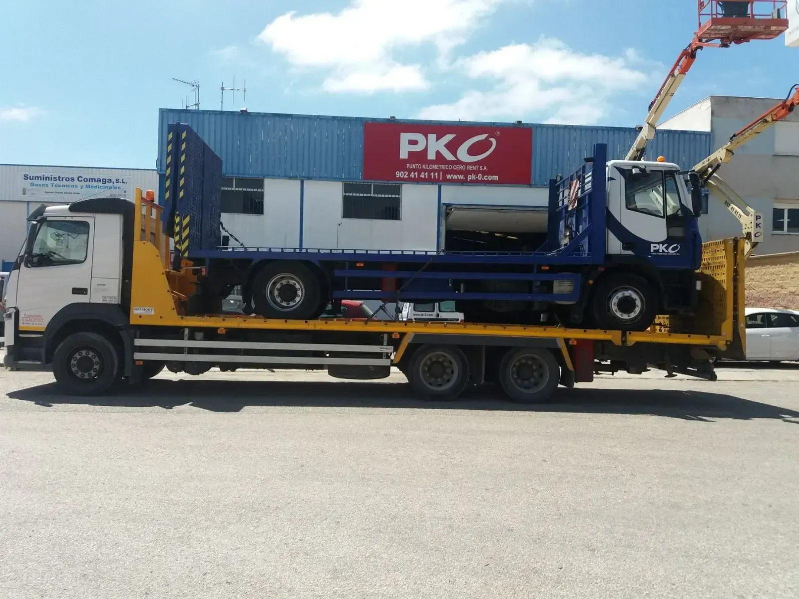 Camión que transporta un contenedor azul con el letrero rojo de PKC en un remolque de plataforma bajo un cielo soleado.