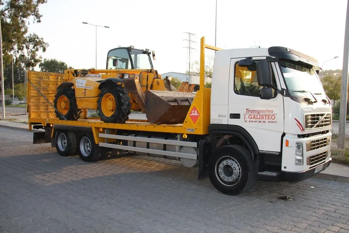 Bulldozer amarillo sobre un camión de plataforma con cabina blanca, estacionado al aire libre al anochecer.