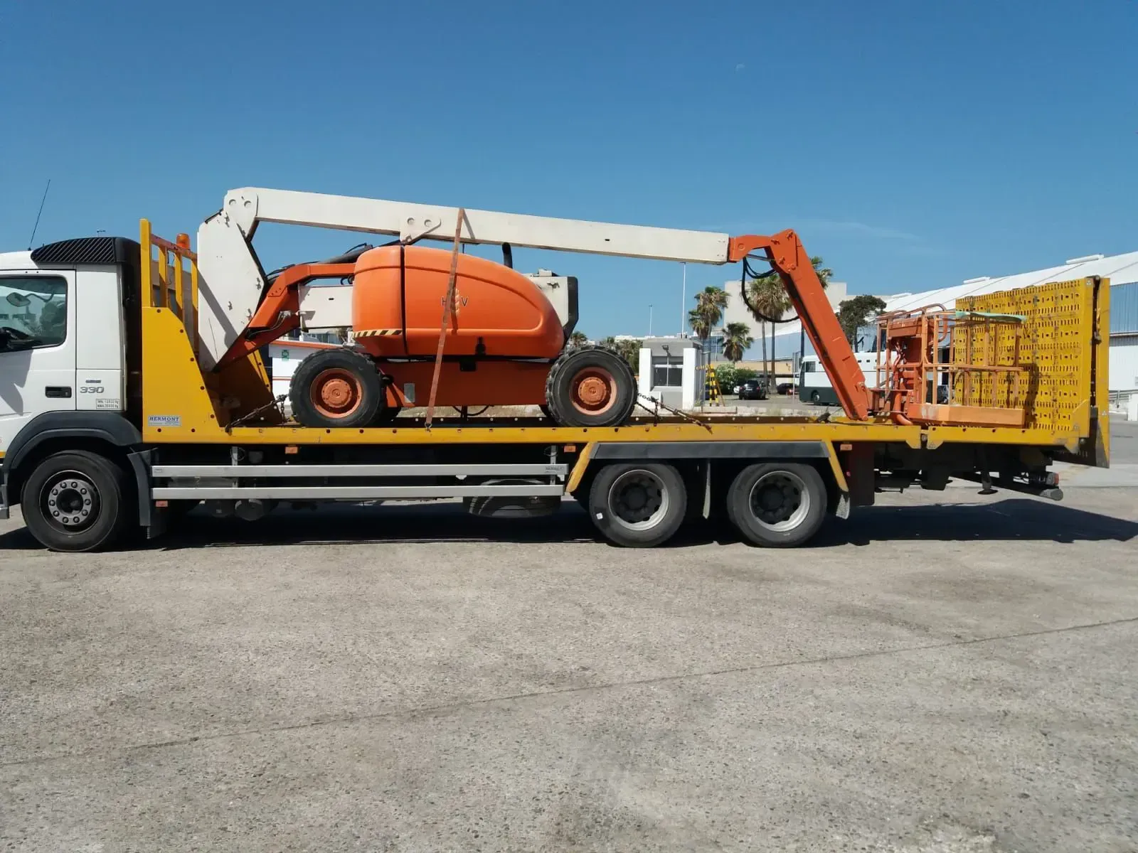 Camión de plataforma que transporta una plataforma elevadora naranja en una obra de construcción en un día soleado.