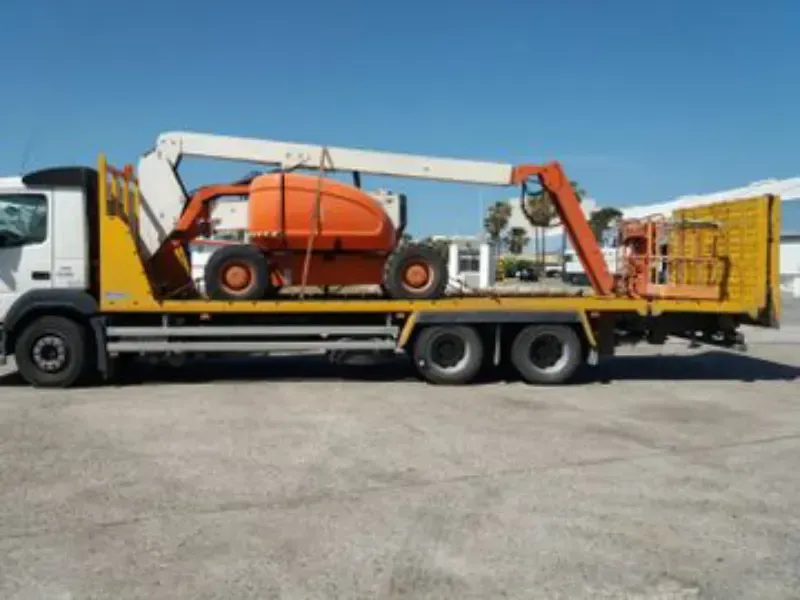 Una plataforma elevadora naranja montada sobre un camión de plataforma en un estacionamiento, con una puerta amarilla detrás.