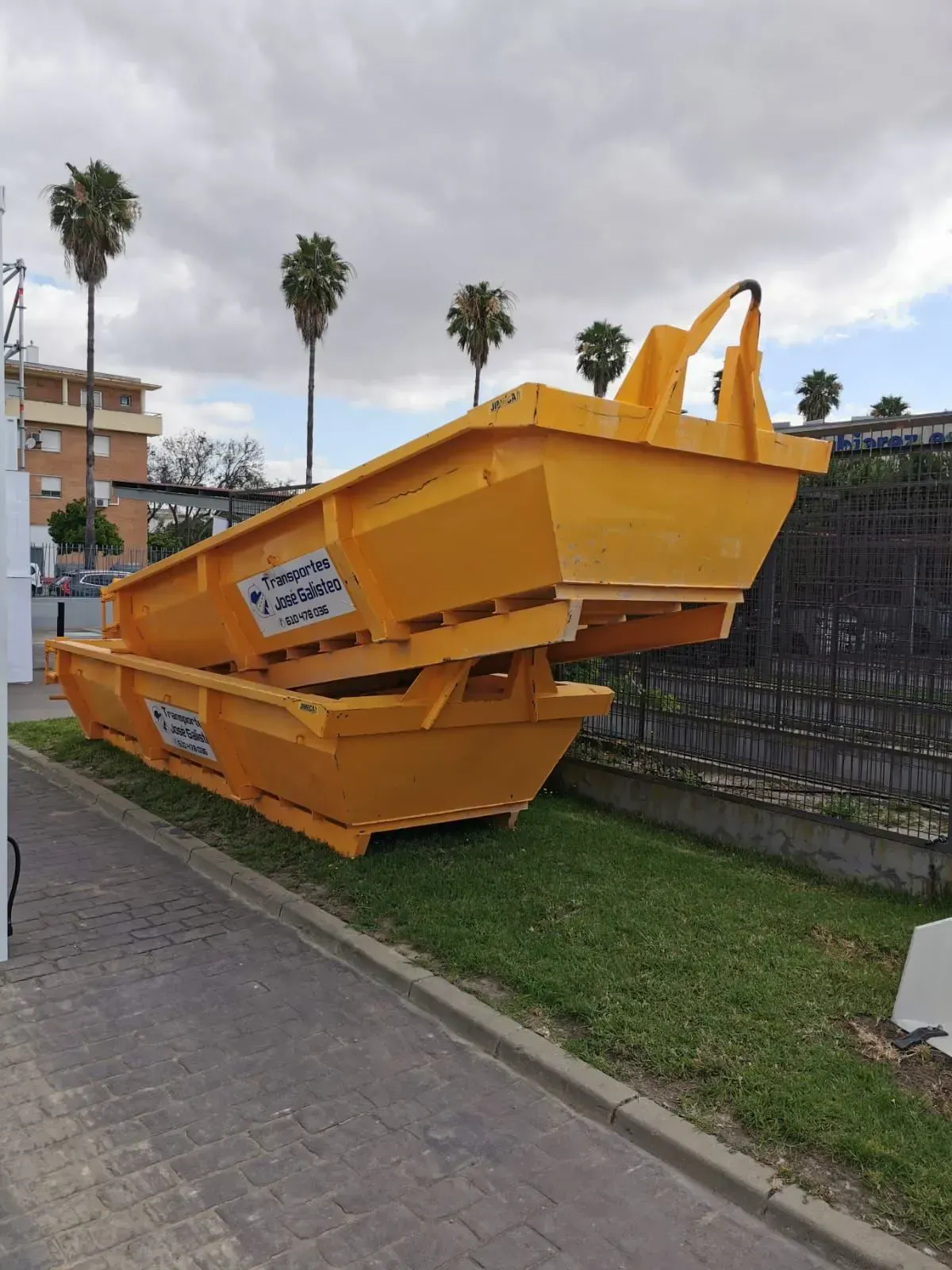 Un gran contenedor de basura metálico de color naranja se encuentra en una acera junto a un muro cubierto de césped