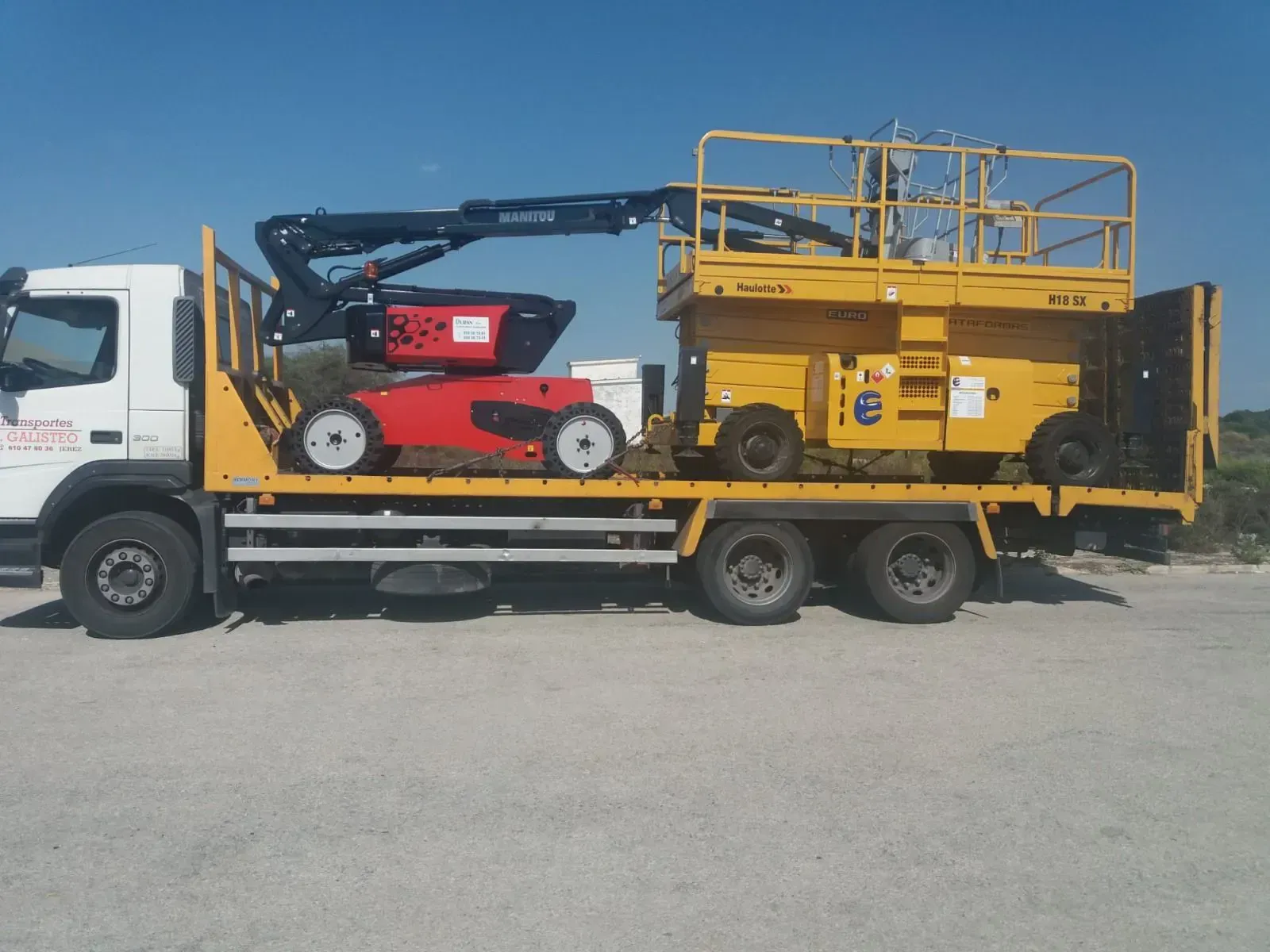 Camión de plataforma que transporta una grúa elevadora roja y una plataforma elevadora amarilla en un terreno de grava.