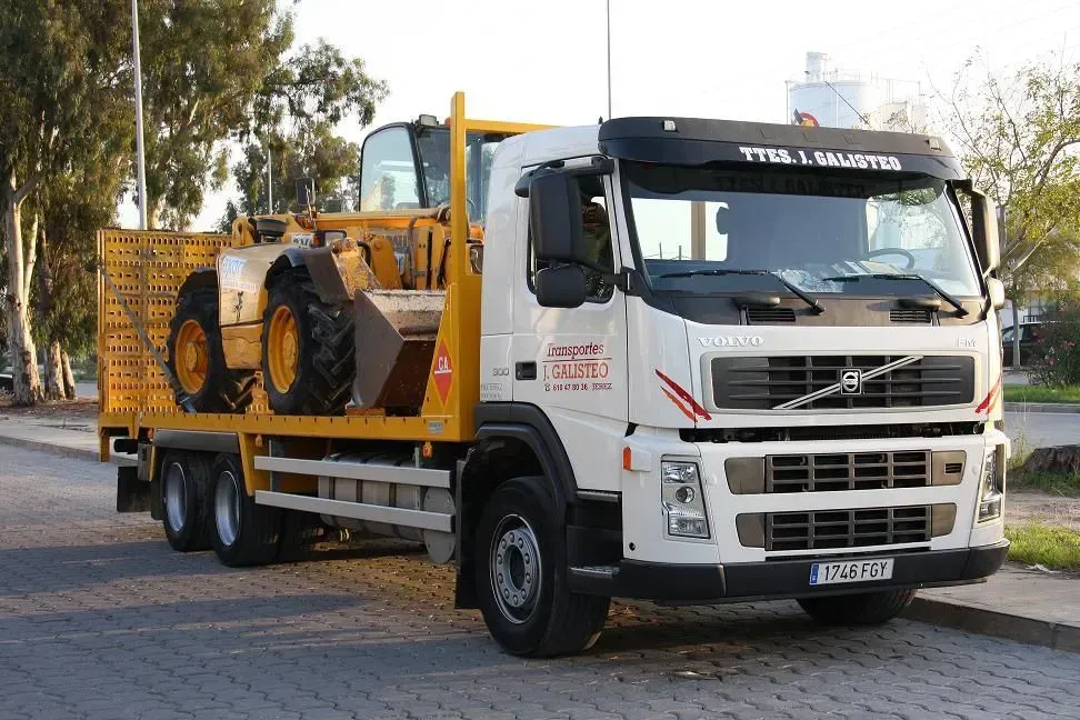 Camión Volvo blanco de plataforma plana que transporta una carretilla elevadora amarilla en una calle exterior