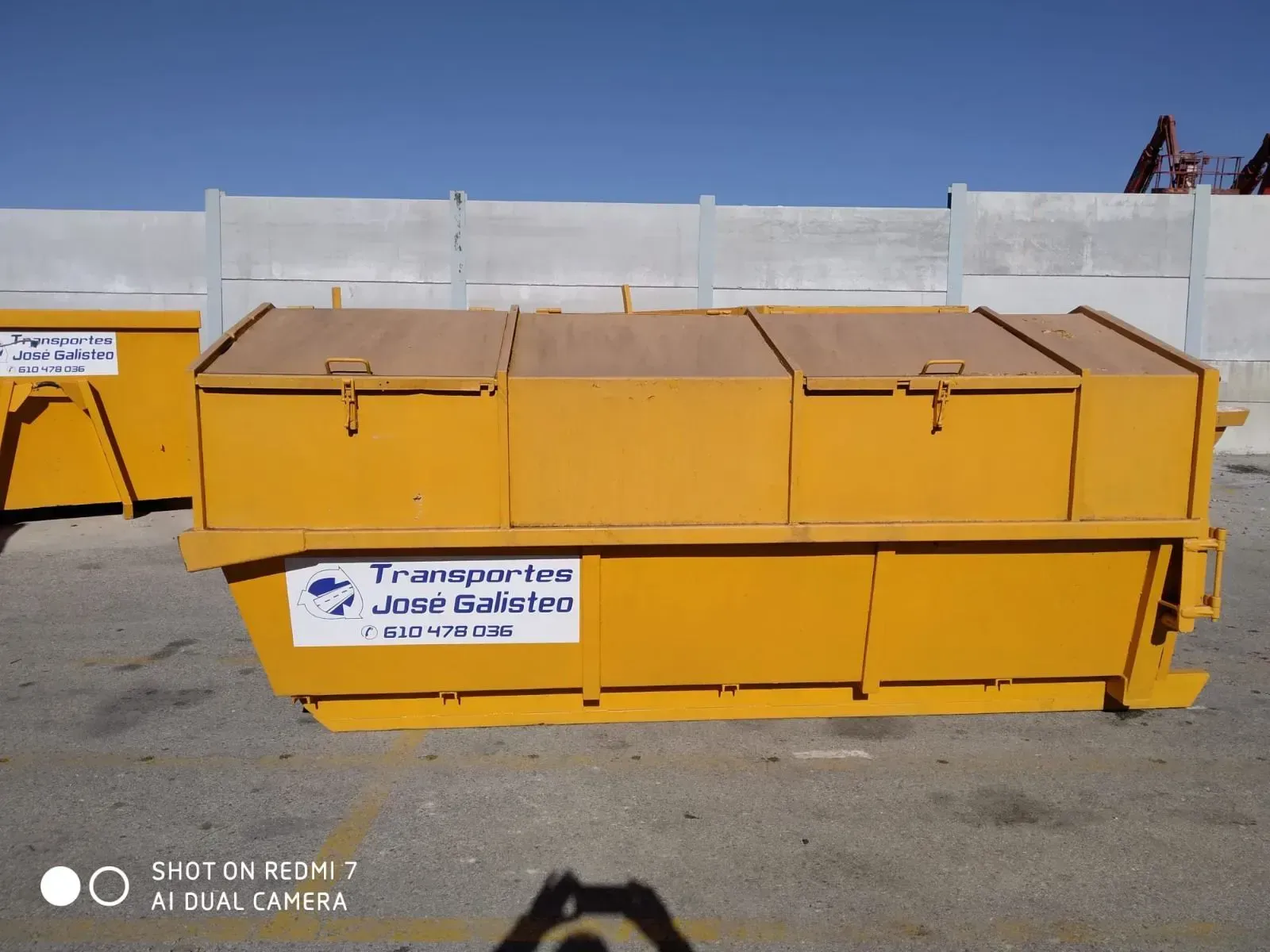 Contenedor industrial amarillo con etiqueta en español en un solar soleado.